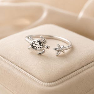Può includere: Un anello aperto color argento con un ciondolo a forma di tartaruga marina su un'estremità e una piccola stella marina sull'altra. La tartaruga presenta un motivo dettagliato sul guscio, mentre la stella marina ha una superficie ruvida.