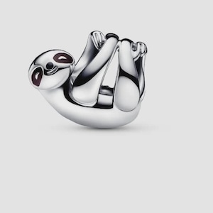 May include: A polished silver charm shaped like a sloth, featuring a smooth, rounded body and limbs curled inward. The sloth has dark, recessed enamel detailing around the eyes and a small, simple etched line for a mouth, creating a minimalist appearance.