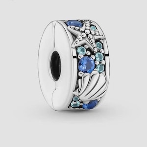 May include: A silver charm bead featuring a nautical theme with a raised starfish and seashell design. The surface is embellished with multiple round, faceted blue and light blue crystals set into the metal, creating a sparkling ocean-inspired look.