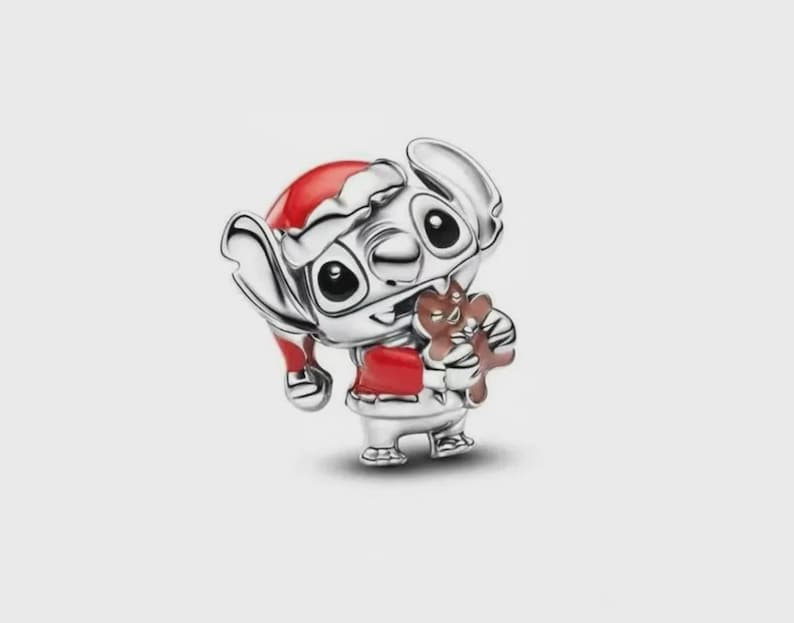 May include: A silver charm featuring a character with large ears wearing a red and white Santa hat and a red jacket, holding a brown gingerbread cookie. The metallic finish is polished and reflective, highlighting the detailed facial features.
