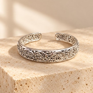May include: A silver-toned open cuff bracelet featuring an intricate, continuous filigree pattern of swirling, interconnected scrollwork. The metal has a polished finish with darkened recessed areas that highlight the detailed relief design.
