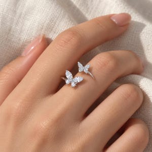 Pode incluir: Um anel aberto de tom prateado com dois motivos de borboletas. A borboleta maior é adornada com pedras transparentes de corte marquise, enquanto a menor é decorada com pequenas pedras redondas e transparentes que brilham.
