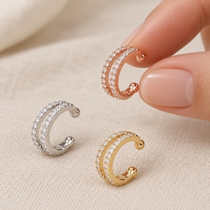 May include: Three double-band ear cuffs, each featuring two rows of small, clear, sparkling stones. One ear cuff is rose gold-toned, one is silver-toned, and one is gold-toned. Each piece has a C-shaped design with rounded ends for secure placement.