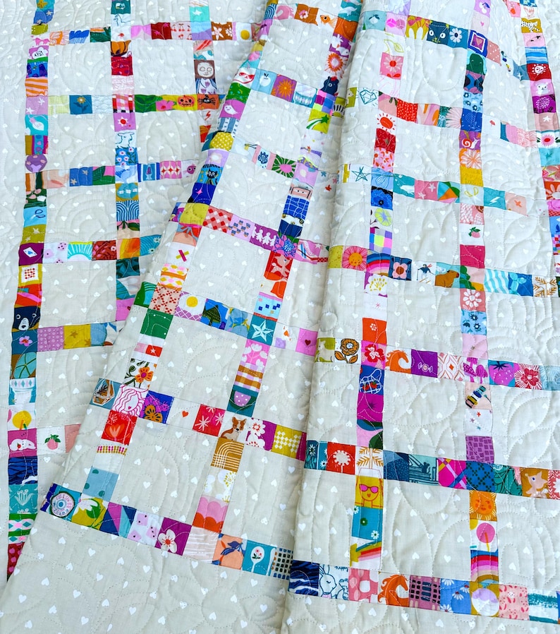 Pu&ograve; includere: Una trapunta patchwork caratterizzata da una griglia di quadrati di tessuto colorati e fantasia, separati da strisce di tessuto color crema. Il tessuto color crema &egrave; decorato con un motivo ripetuto di piccoli cuori bianchi e cuciture trapuntate.