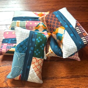 Peut inclure: Une collection de petites pochettes rectangulaires en tissu pr&eacute;sentant un motif patchwork avec divers designs g&eacute;om&eacute;triques dans des tons de bleu sarcelle, orange, rose et blanc. Chaque pochette a une bande centrale en tissu bleu sarcelle.
