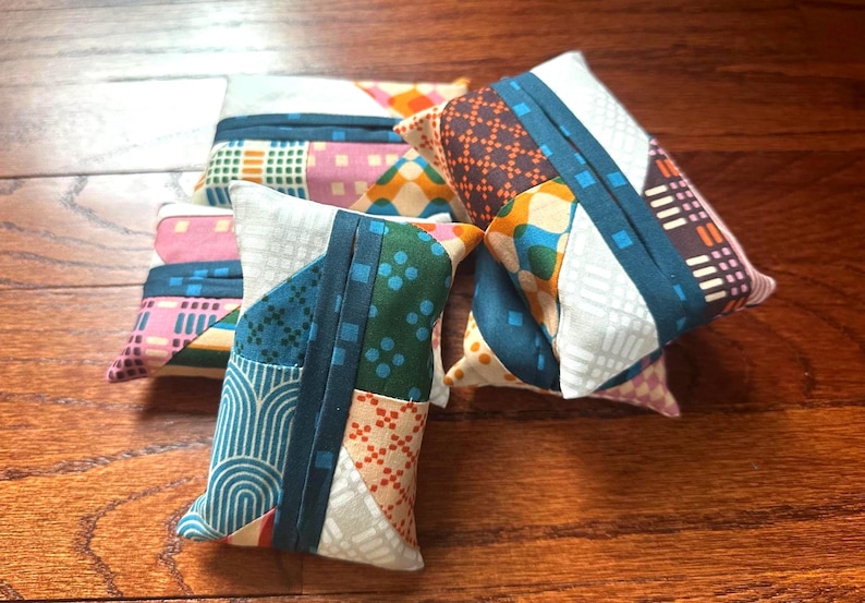 Peut inclure: Une collection de petites pochettes rectangulaires en tissu pr&eacute;sentant un motif patchwork avec divers designs g&eacute;om&eacute;triques dans des tons de bleu sarcelle, orange, rose et blanc. Chaque pochette a une bande centrale en tissu bleu sarcelle.