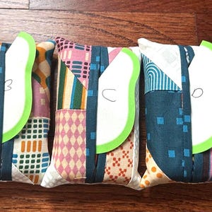 Peut inclure: Cinq pochettes rectangulaires en tissu avec des motifs en patchwork de diff&eacute;rentes couleurs. Chaque pochette poss&egrave;de une sangle verticale bleu sarcelle fonc&eacute; et une &eacute;tiquette verte en forme de feuille marqu&eacute;e des lettres A, B, C, D et E.
