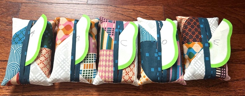 Peut inclure: Cinq pochettes rectangulaires en tissu avec des motifs en patchwork de diff&eacute;rentes couleurs. Chaque pochette poss&egrave;de une sangle verticale bleu sarcelle fonc&eacute; et une &eacute;tiquette verte en forme de feuille marqu&eacute;e des lettres A, B, C, D et E.