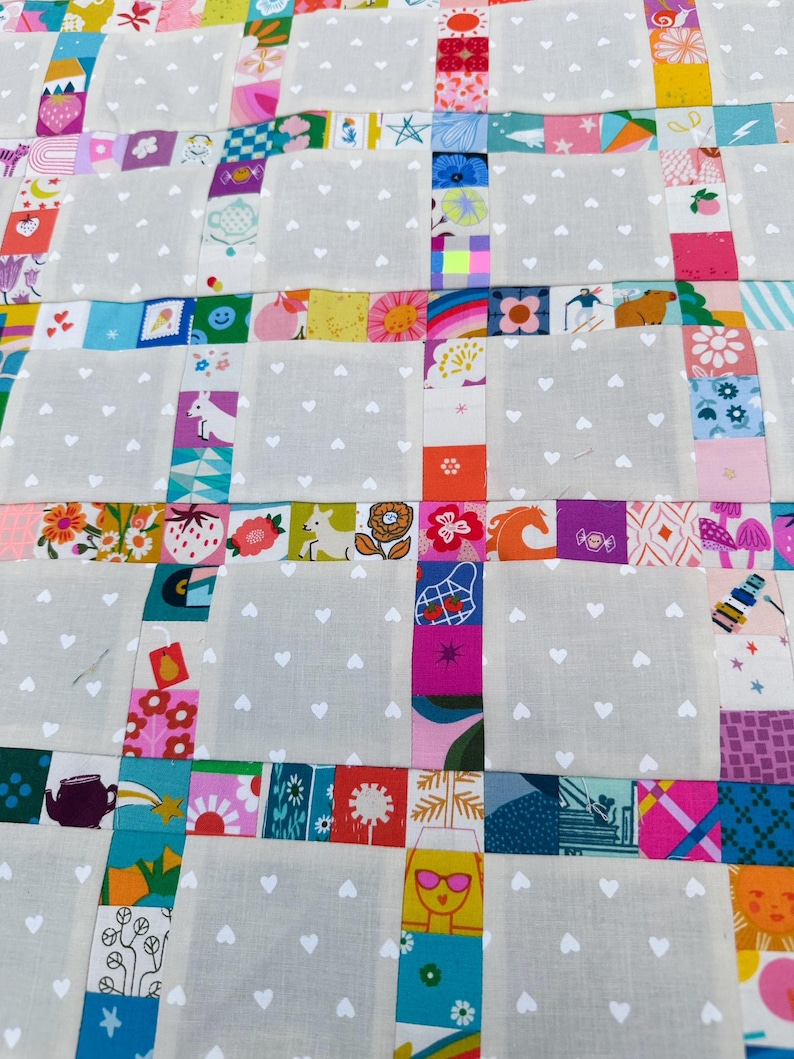Pu&ograve; includere: Una trapunta patchwork con blocchi alternati di tessuto grigio chiaro con un motivo a cuori bianchi e strisce di quadrati di tessuto colorati. I quadrati colorati mostrano varie illustrazioni, tra cui fragole, fiori, animali e forme geometriche.