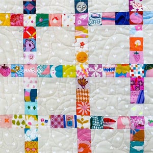 Pu&ograve; includere: Una trapunta patchwork caratterizzata da una griglia di quadrati di tessuto colorati e stampati, separati da blocchi di tessuto beige chiaro con una stampa a cuori bianchi. I quadrati mostrano varie illustrazioni, tra cui una tazza di caff&egrave; e fragole.