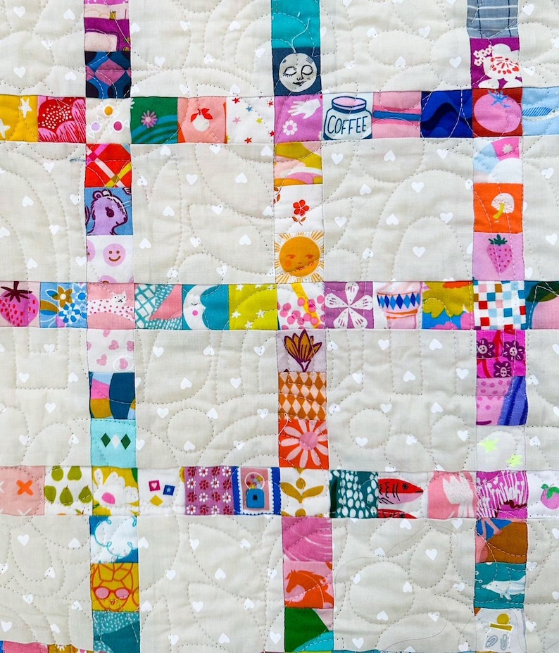 Pu&ograve; includere: Una trapunta patchwork caratterizzata da una griglia di quadrati di tessuto colorati e stampati, separati da blocchi di tessuto beige chiaro con una stampa a cuori bianchi. I quadrati mostrano varie illustrazioni, tra cui una tazza di caff&egrave; e fragole.