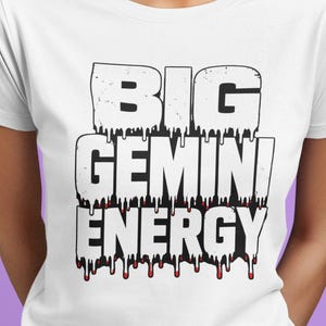 Könnte beinhalten: Ein weißes Kurzarm-T-Shirt mit dem schwarzen Schriftzug BIG GEMINI ENERGY in einer tropfenden, abgenutzten Schriftart. Die Buchstaben haben einen flüssigen Effekt mit kleinen roten Akzenten an den Tropfspitzen, zentriert auf der Brust.
