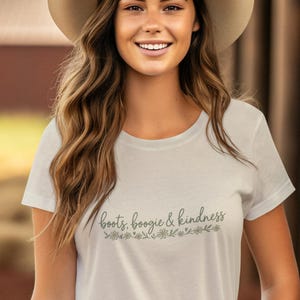 May include: A white short-sleeved t-shirt featuring the text "boots, boogie & kindness" in a cursive font above a small floral graphic. The shirt is paired with a light-colored wide-brimmed hat and olive green pants.