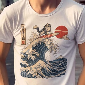 Könnte beinhalten: Ein weißes kurzärmeliges T-Shirt mit einem Grafikdruck einer Katze mit Sonnenbrille, die auf einer großen blauen Welle surft. Das Design enthält eine rote Sonne, einen Berg und den japanischen Text 波に乗る猫, was surfende Katze bedeutet.