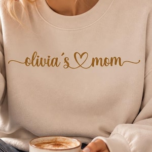 Könnte beinhalten: Ein cremefarbenes Sweatshirt mit Rundhalsausschnitt und dem Schriftzug "olivia's mom" in goldener Schreibschrift, wobei ein Herzsymbol zwischen den Wörtern integriert ist. Das Sweatshirt hat lange Ärmel mit gerippten Bündchen und einen gerippten Halsausschnitt.