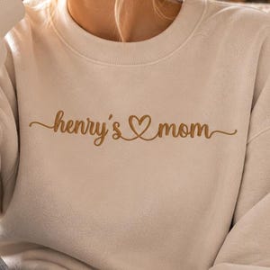 Może przedstawiać: Kremowa bluza z okrągłym dekoltem z napisem "henry's mom" wykonanym brązową kursywą z małym symbolem serca między słowami. Odzież posiada długie rękawy z prążkowanymi mankietami oraz prążkowany dekolt, wykonana z miękkiego materiału.