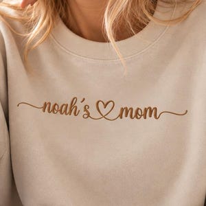 Könnte beinhalten: Ein beigefarbenes Sweatshirt mit Rundhalsausschnitt und dem braun gestickten Text "noah's mom", wobei ein kleines Herzsymbol zwischen den Wörtern integriert ist. Das Kleidungsstück hat lange Ärmel mit Rippbündchen und einen Rippkragen.
