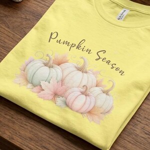 Könnte beinhalten: Ein hellgelbes T-Shirt mit einer Grafik aus mehreren pastellfarbenen Kürbissen und Herbstblättern, wobei der Text "Pumpkin Season" in einer dunkelbraunen Schreibschrift über der Illustration auf der Vorderseite aufgedruckt ist.