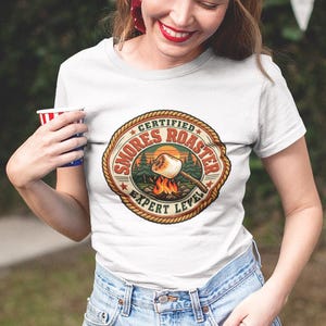 Könnte beinhalten: Ein weißes Kurzarm-T-Shirt mit einer kreisförmigen Grafik mit Seilrand. Das Design zeigt ein Marshmallow an einem Röststab über einem Lagerfeuer, mit dem Text "CERTIFIED SMORES ROASTER EXPERT LEVEL" in kreisförmiger Anordnung.