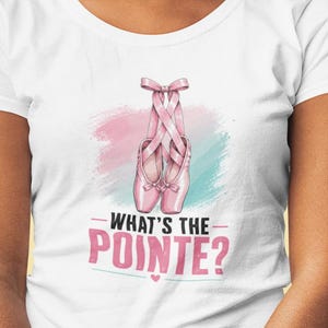Könnte beinhalten: Ein weißes Kurzarm-T-Shirt mit einer Grafik von rosa Ballettschuhen, die mit Bändern gebunden sind. Hinter den Schuhen befindet sich ein weiches rosa und blaues Pinselstrich-Design. Unter der Grafik steht der Text WHAT'S THE POINTE? in Schwarz und Rosa.