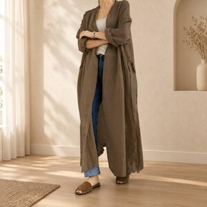 Può includere: Un lungo cardigan color marrone oliva aperto sul davanti con maniche lunghe, indossato sopra una canotta grigio chiaro e jeans blu. L'outfit è completato da sandali marroni, creando un look a strati con una silhouette rilassata e fluida.