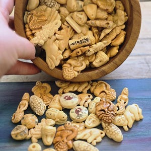 May include: A collection of small, carved, cream-colored beads and charms in various shapes, including animals, faces, and figures, contained within a round wooden bowl and scattered on a surface. One bead features black text markings.