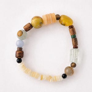 May include: A beaded bracelet featuring a variety of shapes and textures, including round wooden beads, smooth polished stones in shades of yellow, blue, and brown, and cylindrical translucent glass beads arranged in a circular pattern.