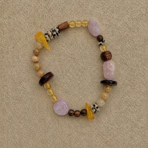 May include: A beaded bracelet featuring a mix of materials including smooth lavender stones, yellow translucent shards, dark brown wooden beads, and small white beads with black spots arranged in a circular pattern on a neutral surface.