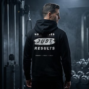 Könnte beinhalten: Schwarzer Kapuzenpullover mit dem weißen Text "NO EXCUSES JUST RESULTS". Der Hoodie hat auch die Wörter "STRENGTH + FOCUS + EVOLVE" unten aufgedruckt. Der Hoodie wird in einem Fitnessstudio getragen.