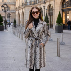 May include: A long-sleeved trench coat featuring a grey and black snakeskin pattern, a notched collar, double-breasted button closure, and a matching tie belt at the waist. The coat is worn over a black turtleneck jumper and dark sunglasses.