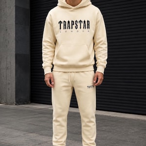 May include: A cream-colored hooded sweatshirt and matching sweatpants set. The hoodie features the black text TRAPSTAR LONDON on the chest and a front kangaroo pocket. The sweatpants have a drawstring waist and the black text TRAPSTAR on the leg.