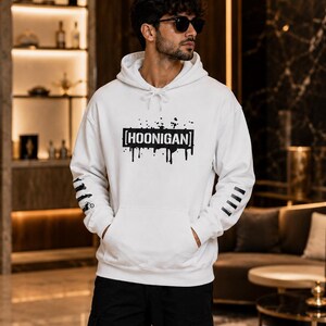 May include: A white hooded sweatshirt featuring a black graphic print on the chest with the text HOONIGAN inside a rectangular box with paint drip effects. The sleeves have black graphic details, and the garment includes a front kangaroo pocket.