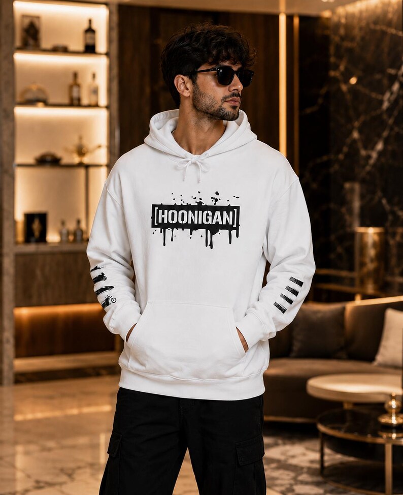 May include: A white hooded sweatshirt featuring a black graphic print on the chest with the text HOONIGAN inside a rectangular box with paint drip effects. The sleeves have black graphic details, and the garment includes a front kangaroo pocket.