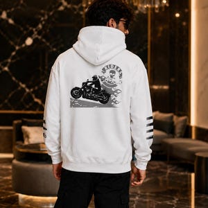 May include: A white hooded sweatshirt featuring a black and white graphic on the back. The design depicts a motorcycle rider, flames, and a circular emblem with a skull and the text RIDERS. The left sleeve has three horizontal black stripes.