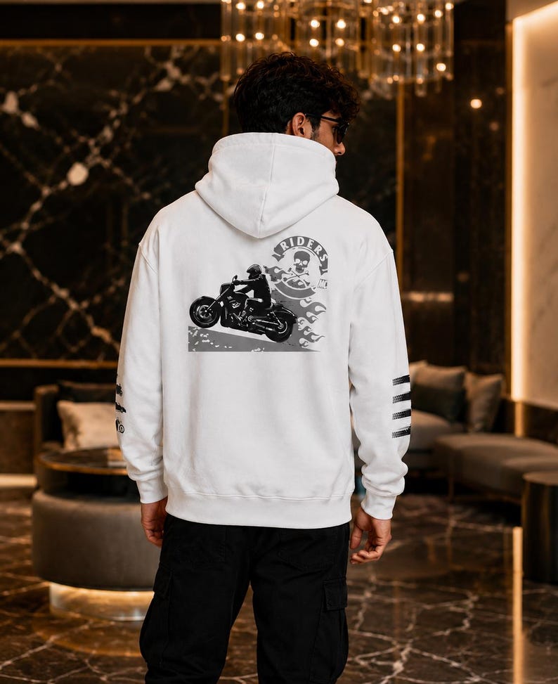 May include: A white hooded sweatshirt featuring a black and white graphic on the back. The design depicts a motorcycle rider, flames, and a circular emblem with a skull and the text RIDERS. The left sleeve has three horizontal black stripes.