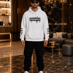 May include: A white hooded sweatshirt featuring a black graphic print with the text HOONIGAN in a rectangular frame with paint drip effects. The sleeves have black horizontal stripes, paired with black cargo trousers and white trainers.
