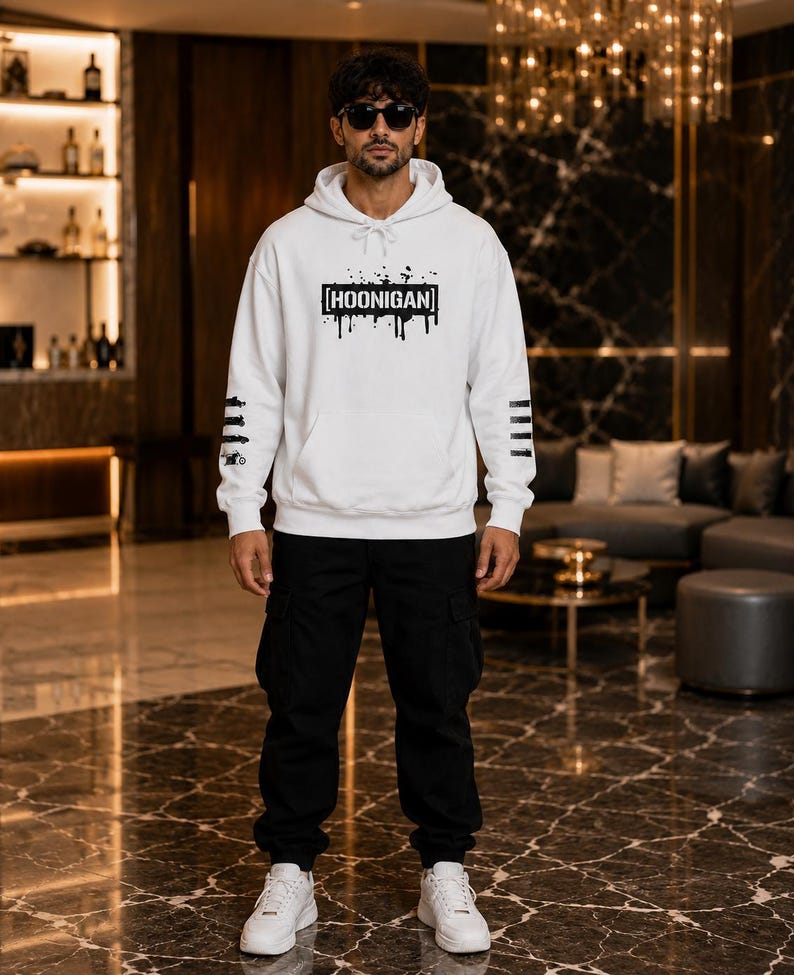 May include: A white hooded sweatshirt featuring a black graphic print with the text HOONIGAN in a rectangular frame with paint drip effects. The sleeves have black horizontal stripes, paired with black cargo trousers and white trainers.