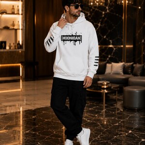 May include: A white hooded sweatshirt featuring a black graphic print on the chest that reads HOONIGAN inside a rectangular box with paint drip effects. The sleeves have black horizontal stripes, and the garment includes a front kangaroo pocket.