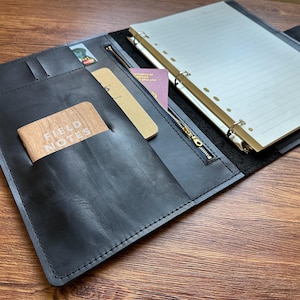 May include: A black leather organiser folio featuring a six-ring binder mechanism with lined paper, a zipped pocket, and multiple interior slots. A tan Field Notes notebook is tucked into a front pocket, alongside a passport and credit cards.