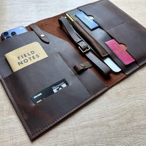 May include: A dark brown leather travel organiser featuring multiple interior pockets and a strap closure. It holds a Field Notes notebook, a smartphone, a pen, a passport, and credit cards. The organiser is constructed with visible stitching.