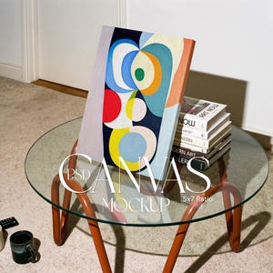 May include: A rectangular canvas print featuring an abstract composition of overlapping circles and shapes in blue, yellow, red, orange, and green. The text PSD CANVAS MOCKUP 5x7 Ratio is overlaid on the image. The canvas is propped against a stack of books.