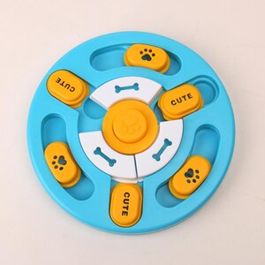 May include: A blue and yellow dog puzzle toy. The toy has a circular design with multiple compartments and sliding pieces. The toy has the word "CUTE" printed on some of the yellow pieces. The toy is designed to stimulate a dog's mind.