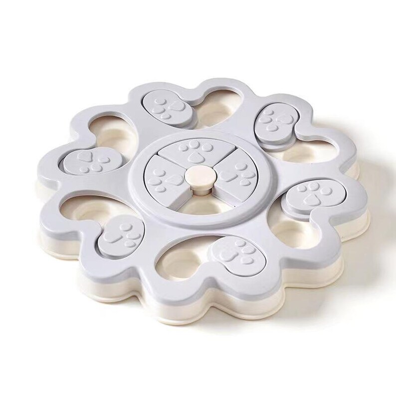 May include: A light grey dog puzzle toy with a flower-like design. The toy features multiple compartments and paw print details. The centre has a rotating section with paw print shapes. The base is white.