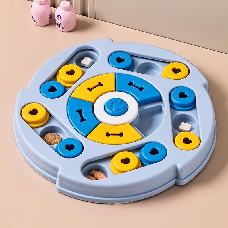 May include: A light blue dog puzzle toy with yellow, blue, and white elements. The toy features compartments and sliding parts for treats. Two pink bottles are visible in the background.