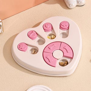 May include: A pink and white paw-shaped dog puzzle toy with various compartments and sliding pieces. The toy features pink bone and heart-shaped covers, designed to hide treats. The toy is designed to stimulate a dog's mind and provide enrichment.