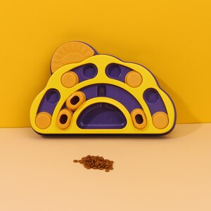 May include: A yellow and purple pet puzzle toy in a paw shape. It features various compartments and sliding elements. A small pile of brown pet food is in front of the toy, indicating its purpose. The background is a solid yellow.