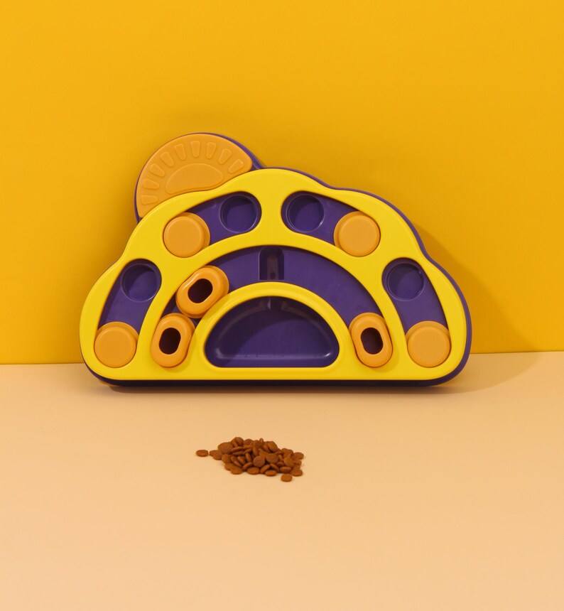 May include: A yellow and purple pet puzzle toy in a paw shape. It features various compartments and sliding elements. A small pile of brown pet food is in front of the toy, indicating its purpose. The background is a solid yellow.