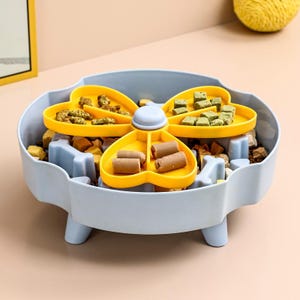 May include: A light blue dog food puzzle bowl with a yellow, four-sectioned, rotating food dispenser. The dispenser holds various dog treats. The bowl has a bone-shaped design and is filled with dog food.
