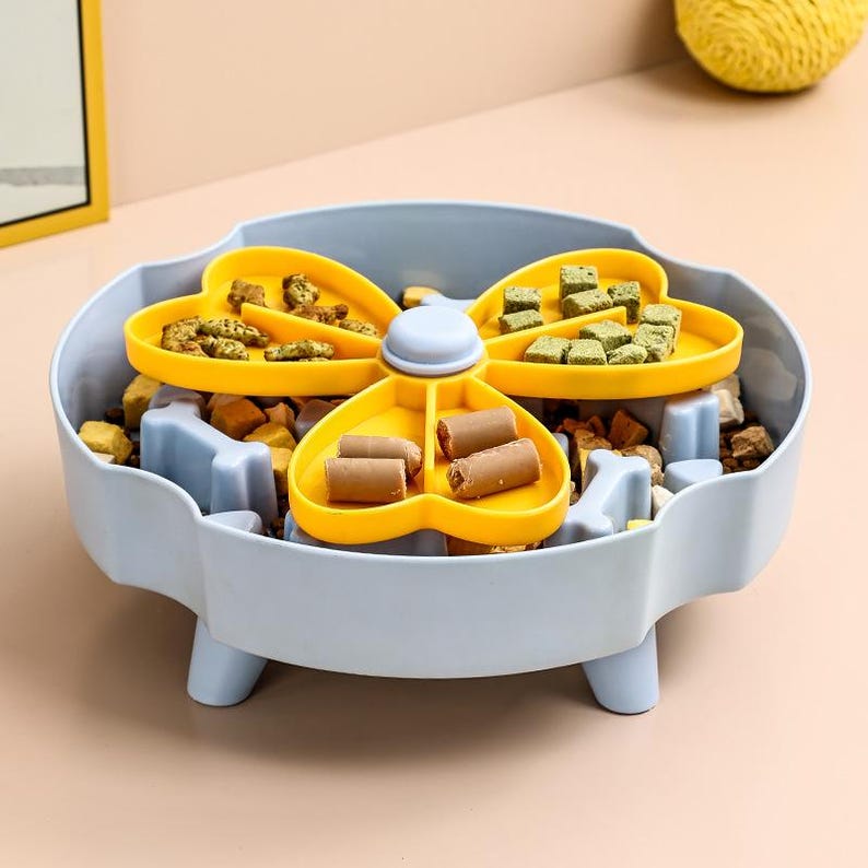 May include: A light blue dog food puzzle bowl with a yellow, four-sectioned, rotating food dispenser. The dispenser holds various dog treats. The bowl has a bone-shaped design and is filled with dog food.