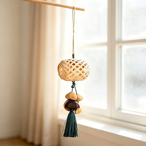 May include: A hanging ornament featuring a light-colored, woven bamboo sphere suspended by a thin cord. Below the sphere, a dark green tassel hangs alongside several smooth, polished wooden beads in varying shades of brown and tan.